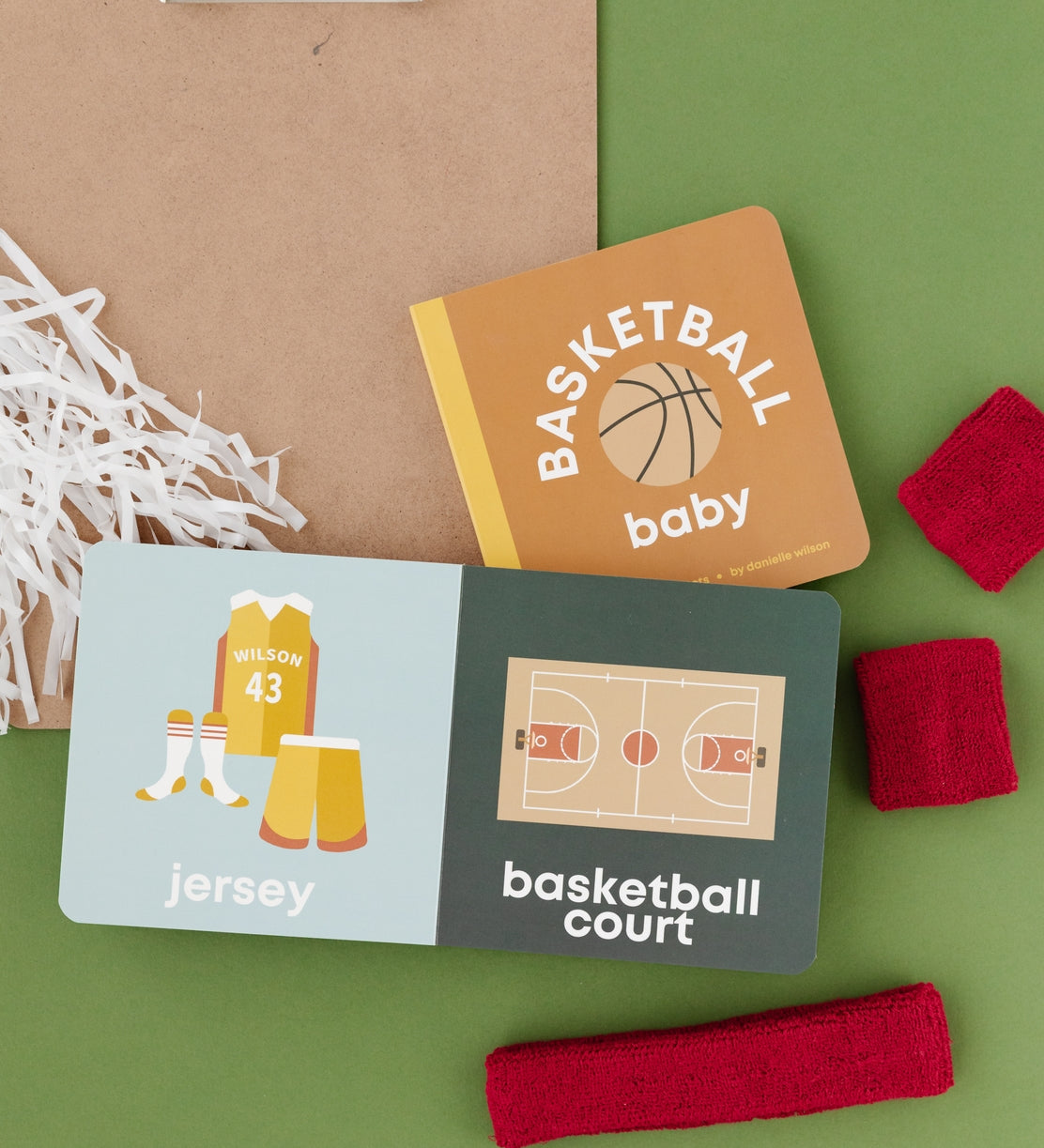 Basketball-themed baby items including a book, cards, and headbands on a green background. Availabe at Be Charmed