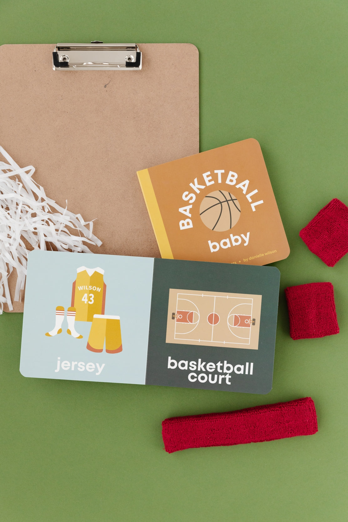 Basketball-themed baby items including a book, cards, and headbands on a green background. Availabe at Be Charmed