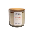 A 16 oz soy candle in a clear glass jar with a light wood lid, featuring a white label that reads 'Lola & Bean Co. Autumn Harvest Pumpkin-Clove Oak'. Available at Be Charmed