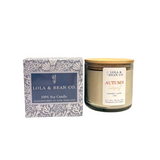 Lola & Bean Co. Autumn Harvest handpoured soy candle in a clear glass jar with a light wood lid, shown next to its patterned blue and white packaging box on a white background. Available at Be Charmed