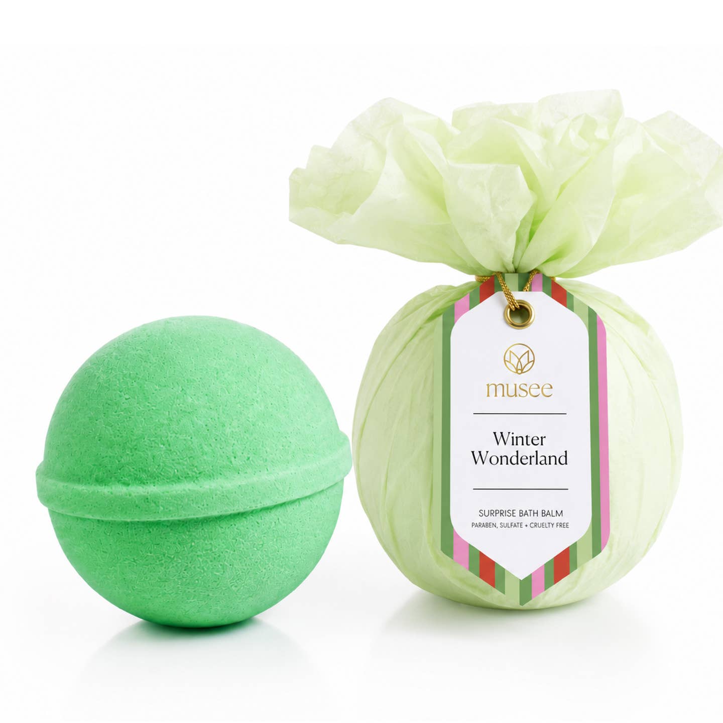 A Musee bath bomb next to its wrapped package, which features a pale green tissue paper flower and a white tag reading ' musee Winter Wonderland'. Available at Be Charmed
