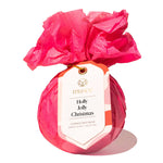 A Musee bath balm wrapped in bright pink tissue paper and tied with a paper hangtag that reads 'musée Holly Jolly Christmas' on a white background. Available at Be Charmed