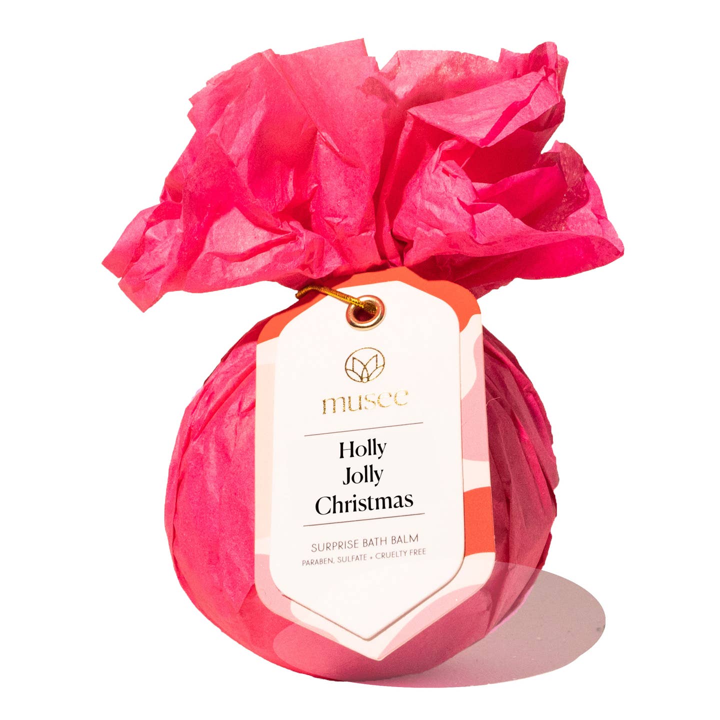 A Musee bath balm wrapped in bright pink tissue paper and tied with a paper hangtag that reads 'musée Holly Jolly Christmas' on a white background. Available at Be Charmed