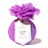 A purple wrapped bath bomb package tied with a string and featuring a white hang tag that reads 'mussee Lavender Haze Surprise Bath Balm,' topped with crinkled purple tissue paper, on a white background. Available at Be Charmed