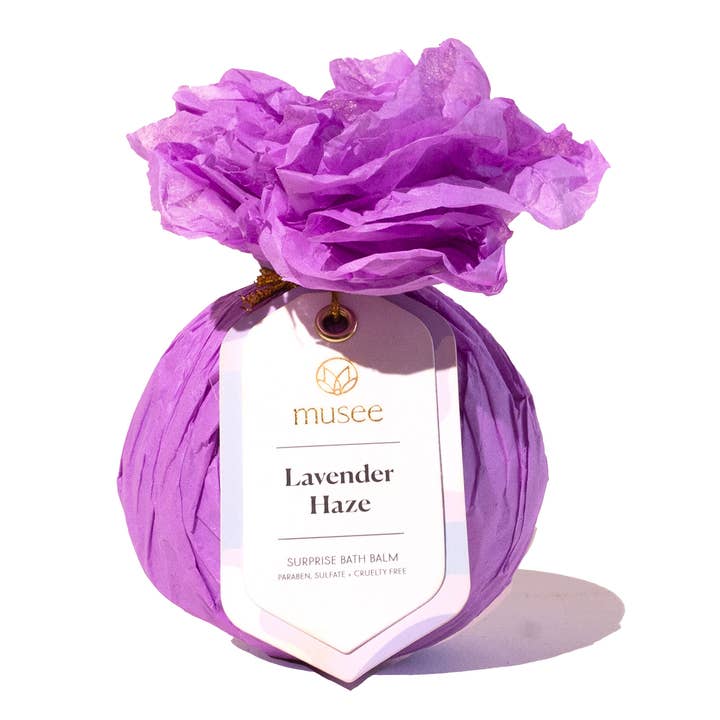 A purple wrapped bath bomb package tied with a string and featuring a white hang tag that reads 'mussee Lavender Haze Surprise Bath Balm,' topped with crinkled purple tissue paper, on a white background. Available at Be Charmed
