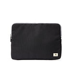 A black fabric laptop sleeve with a small beige brand patch on the bottom right corner, shown against a plain white background. Available at Be Charmed