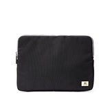 A black fabric laptop sleeve with a small beige brand patch on the bottom right corner, shown against a plain white background. Available at Be Charmed