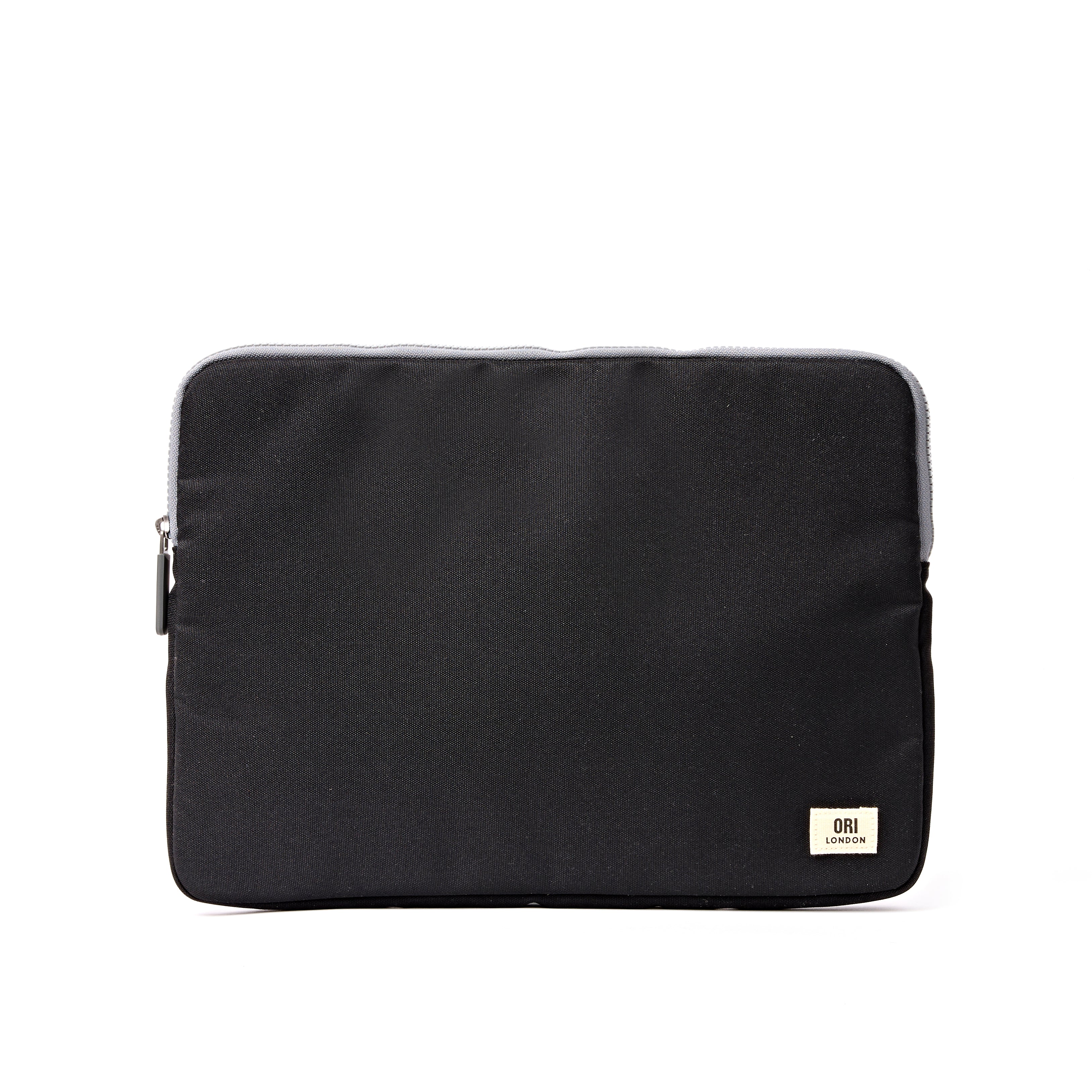 A black fabric laptop sleeve with a small beige brand patch on the bottom right corner, shown against a plain white background. Available at Be Charmed