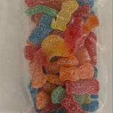 A clear plastic bag filled with assorted colorful sour gummy candies, photographed from above on a plain white surface. Available at Be Charmed