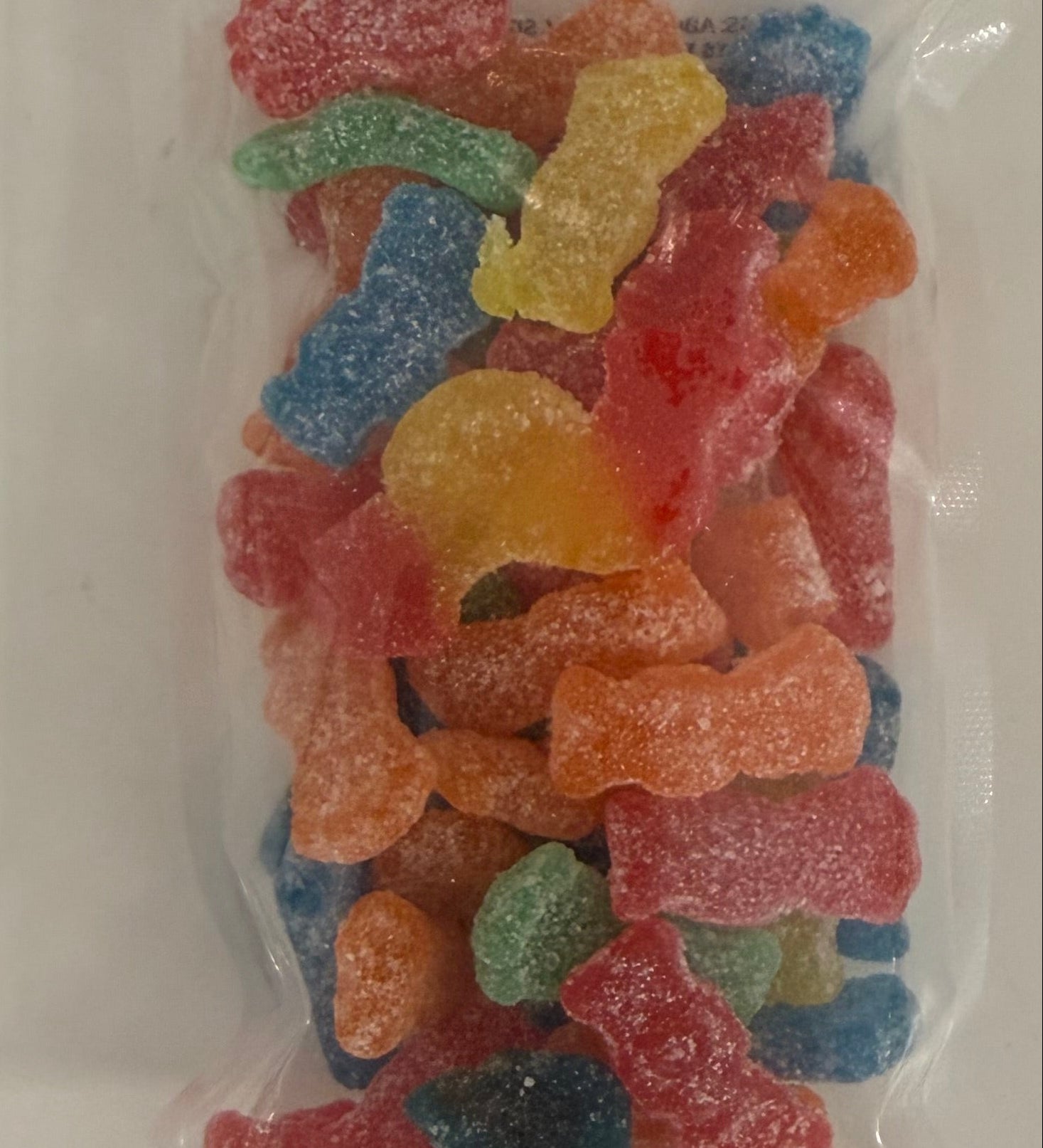 A clear plastic bag filled with assorted colorful sour gummy candies, photographed from above on a plain white surface. Available at Be Charmed