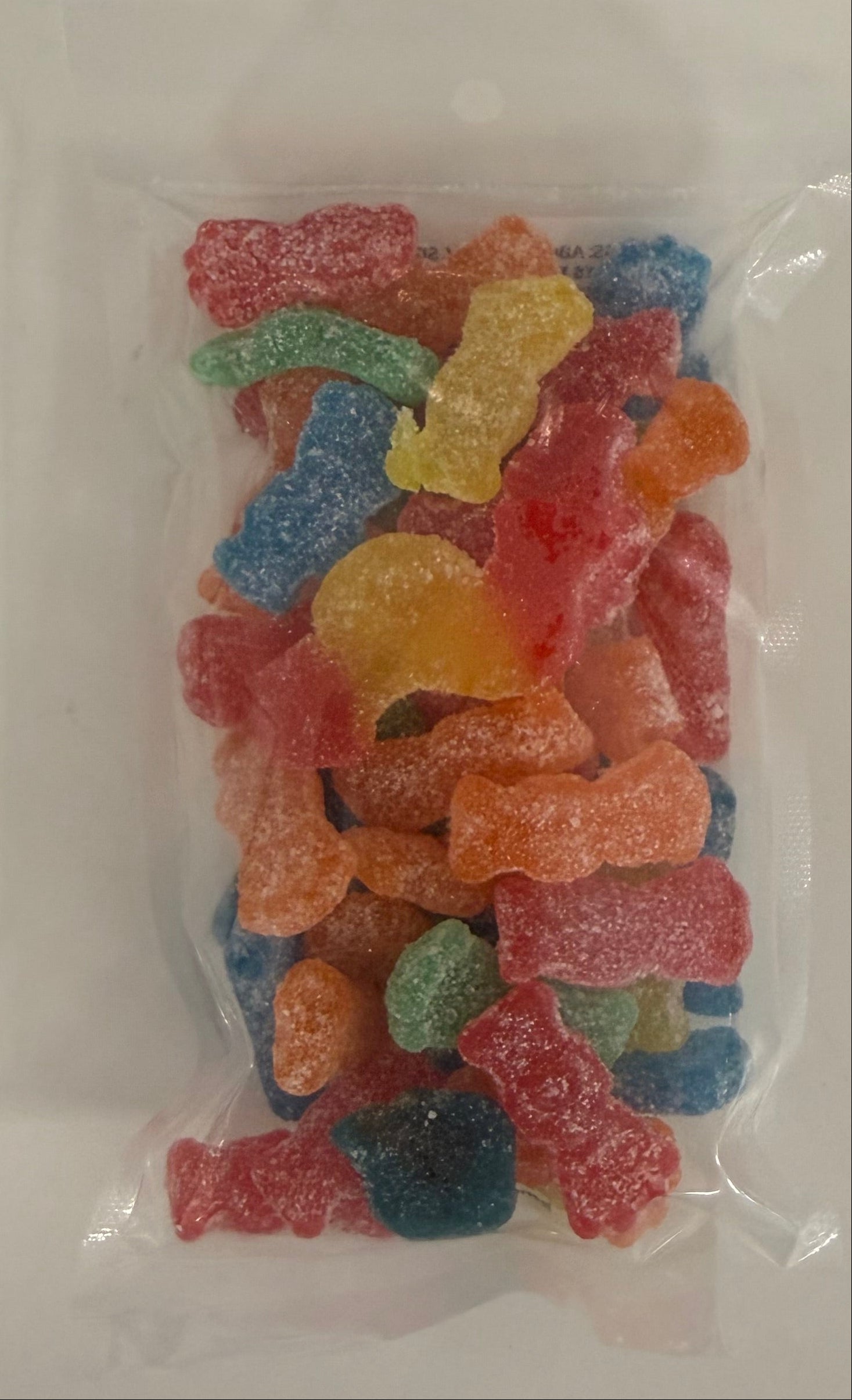 A clear plastic bag filled with assorted colorful sour gummy candies, photographed from above on a plain white surface. Available at Be Charmed