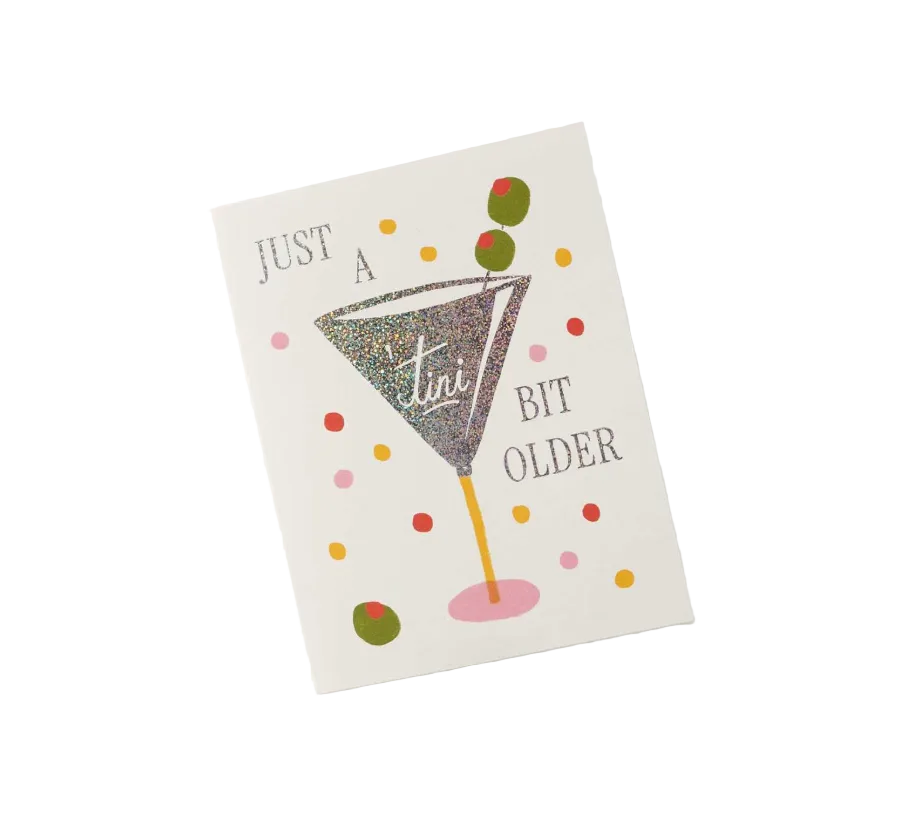 A square greeting card with a white background featuring a glittery martini glass illustration and the text 'JUST A TINI BIT OLDER' in gray, surrounded by colorful confetti dots. Available at Be Charmed