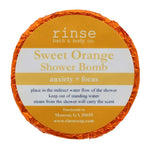 A circular blue shower bomb packaging with a label that reads 'Sweet Orange Shower Bomb' from Rinse Bath & Body Co. Available at Be Charmed