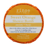 A circular blue shower bomb packaging with a label that reads 'Sweet Orange Shower Bomb' from Rinse Bath & Body Co. Available at Be Charmed