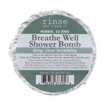A round foil-wrapped bath bomb package labeled 'rinse bath & body co.' and 'Breathe Well Shower Bomb' on a plain white background. Available at Be Charmed