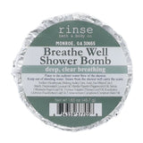 A round foil-wrapped bath bomb package labeled 'rinse bath & body co.' and 'Breathe Well Shower Bomb' on a plain white background. Available at Be Charmed