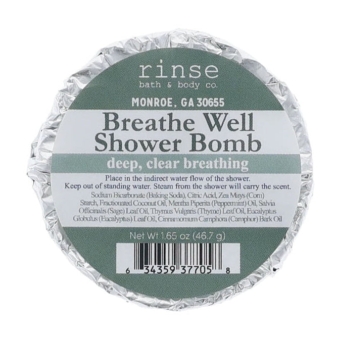A round foil-wrapped bath bomb package labeled 'rinse bath & body co.' and 'Breathe Well Shower Bomb' on a plain white background. Available at Be Charmed
