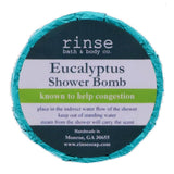 A circular blue shower bomb packaging with a label that reads 'Eucalyptus Shower Bomb' from Rinse Bath & Body Co. Available at Be Charmed