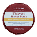 A circular blue shower bomb packaging with a label that reads 'Thievery Shower Bomb' from Rinse Bath & Body Co. Available at Be Charmed