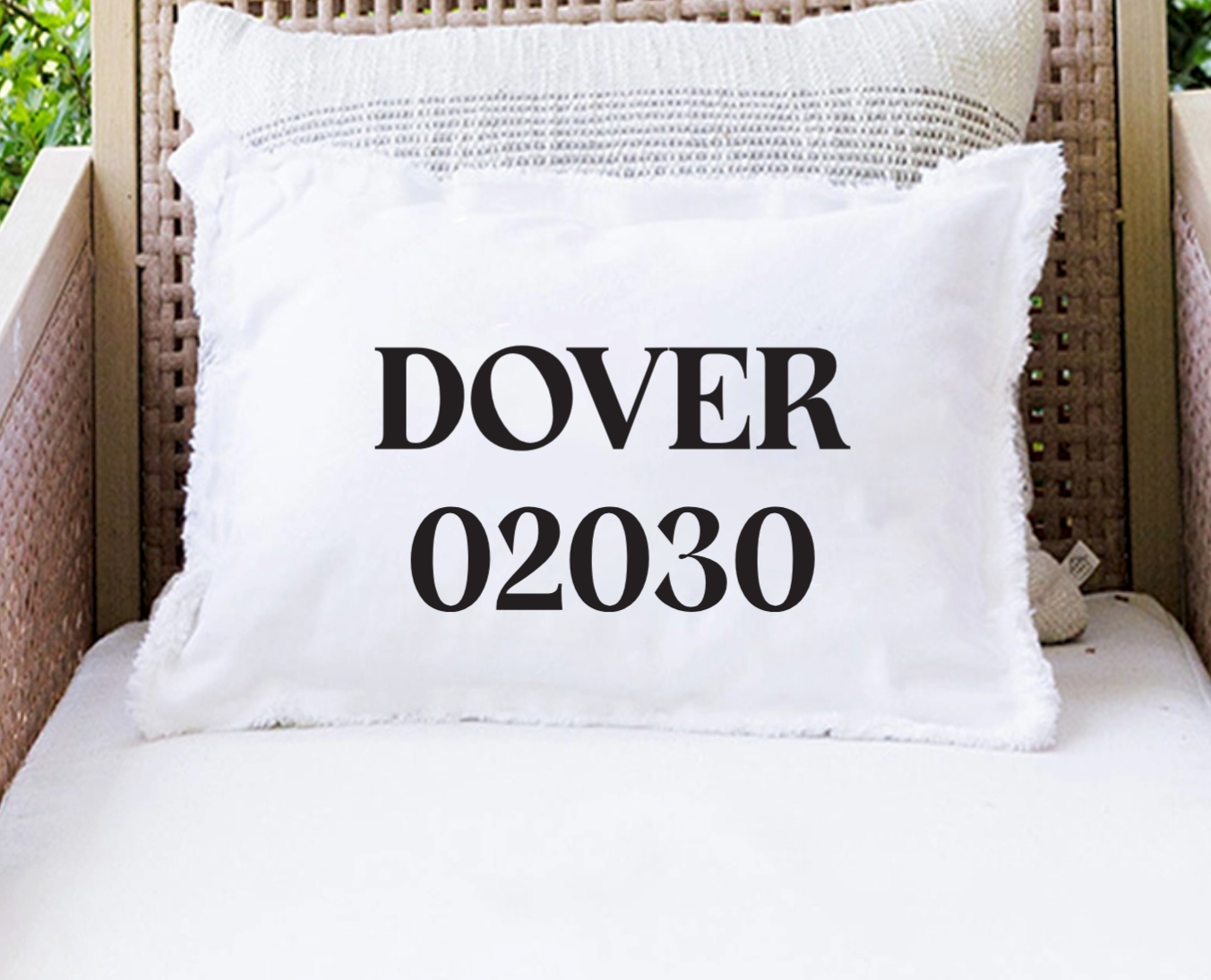 A white throw pillow with black text reading 'DOVER 02030' sits on a white cushion in a wicker chair, with a second textured pillow behind it. Available at BE Charmed