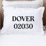 A white throw pillow with black text reading 'DOVER 02030' sits on a white cushion in a wicker chair, with a second textured pillow behind it. Available at BE Charmed