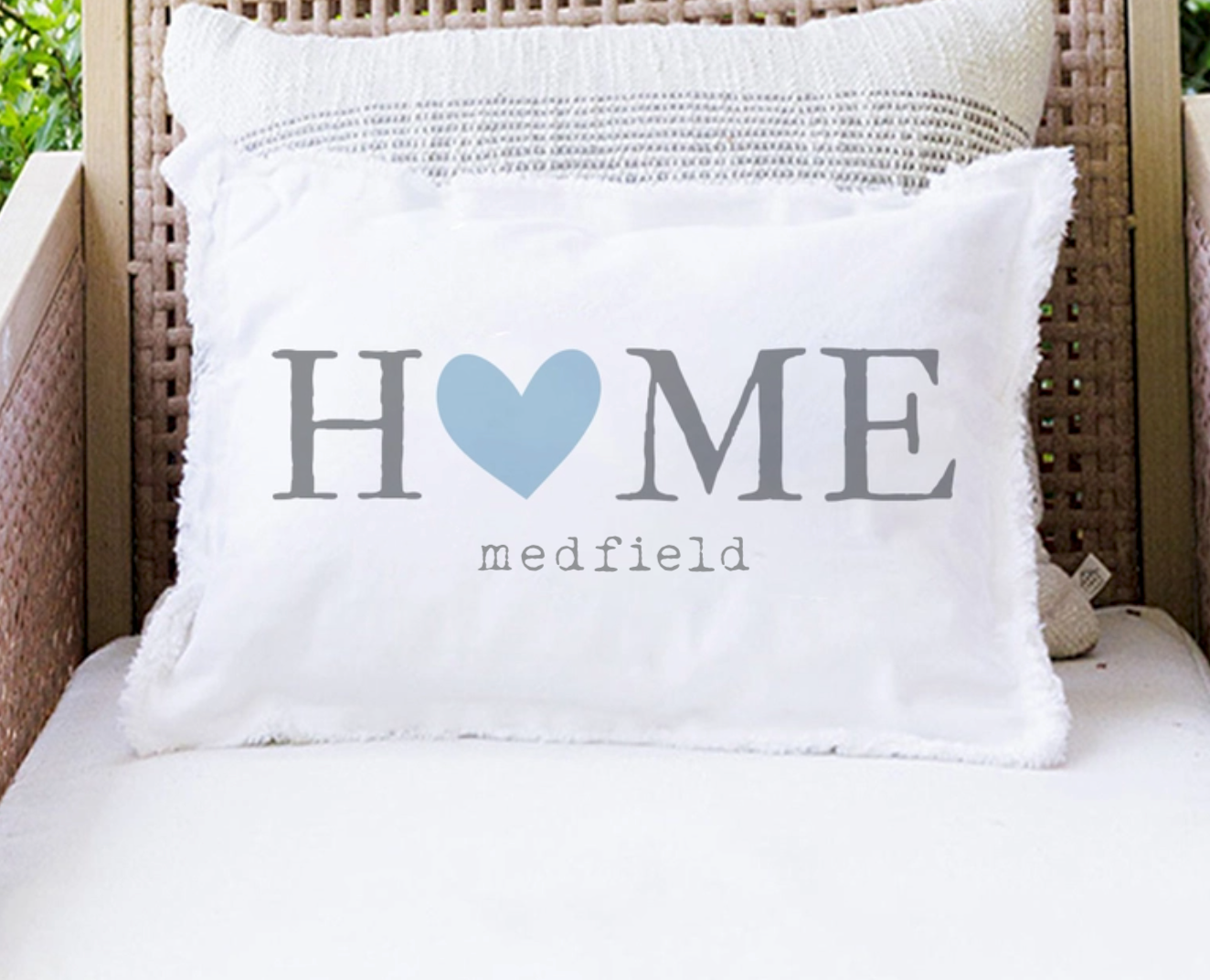 A white rectangular throw pillow with fringed edges sits on a white cushion in a wicker chair. The pillow is printed with the word 'HOME' in gray, where the 'O' is a light blue heart, and 'medfield' in small gray text below. Available at Be Charmed