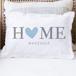 A white rectangular throw pillow with fringed edges sits on a white cushion in a wicker chair. The pillow is printed with the word 'HOME' in gray, where the 'O' is a light blue heart, and 'medfield' in small gray text below. Available at Be Charmed