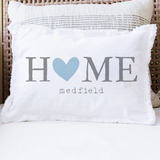 A white rectangular throw pillow with fringed edges sits on a white cushion in a wicker chair. The pillow is printed with the word 'HOME' in gray, where the 'O' is a light blue heart, and 'medfield' in small gray text below. Available at Be Charmed