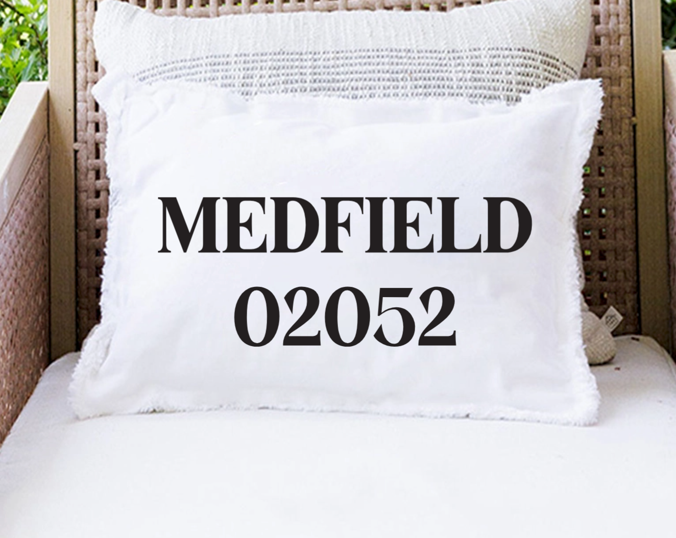 A white rectangular throw pillow with black text reading 'MEDFIELD 02052' sits on a white cushion in front of a woven wicker chair. Available at Be Charmed
