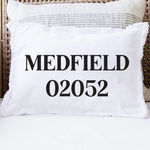 A white rectangular throw pillow with black text reading 'MEDFIELD 02052' sits on a white cushion in front of a woven wicker chair. Available at Be Charmed