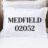 A white rectangular throw pillow with black text reading 'MEDFIELD 02052' sits on a white cushion in front of a woven wicker chair. Available at Be Charmed