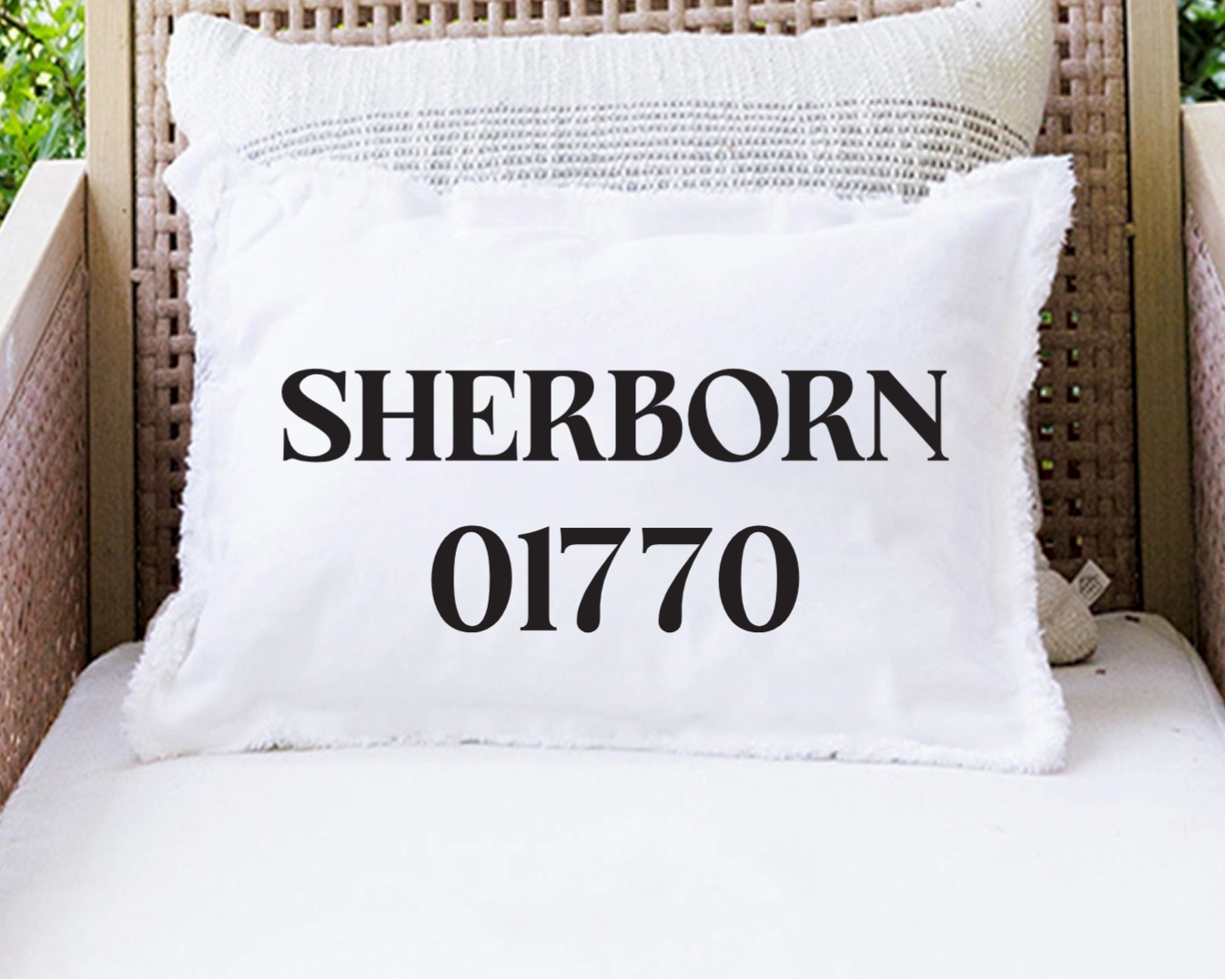 A white decorative throw pillow with fringed edges printed with the text 'SHERBORN 01770' in bold black lettering, resting on a white cushion in a wicker outdoor chair. Available at Be Charmed