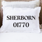 A white decorative throw pillow with fringed edges printed with the text 'SHERBORN 01770' in bold black lettering, resting on a white cushion in a wicker outdoor chair. Available at Be Charmed