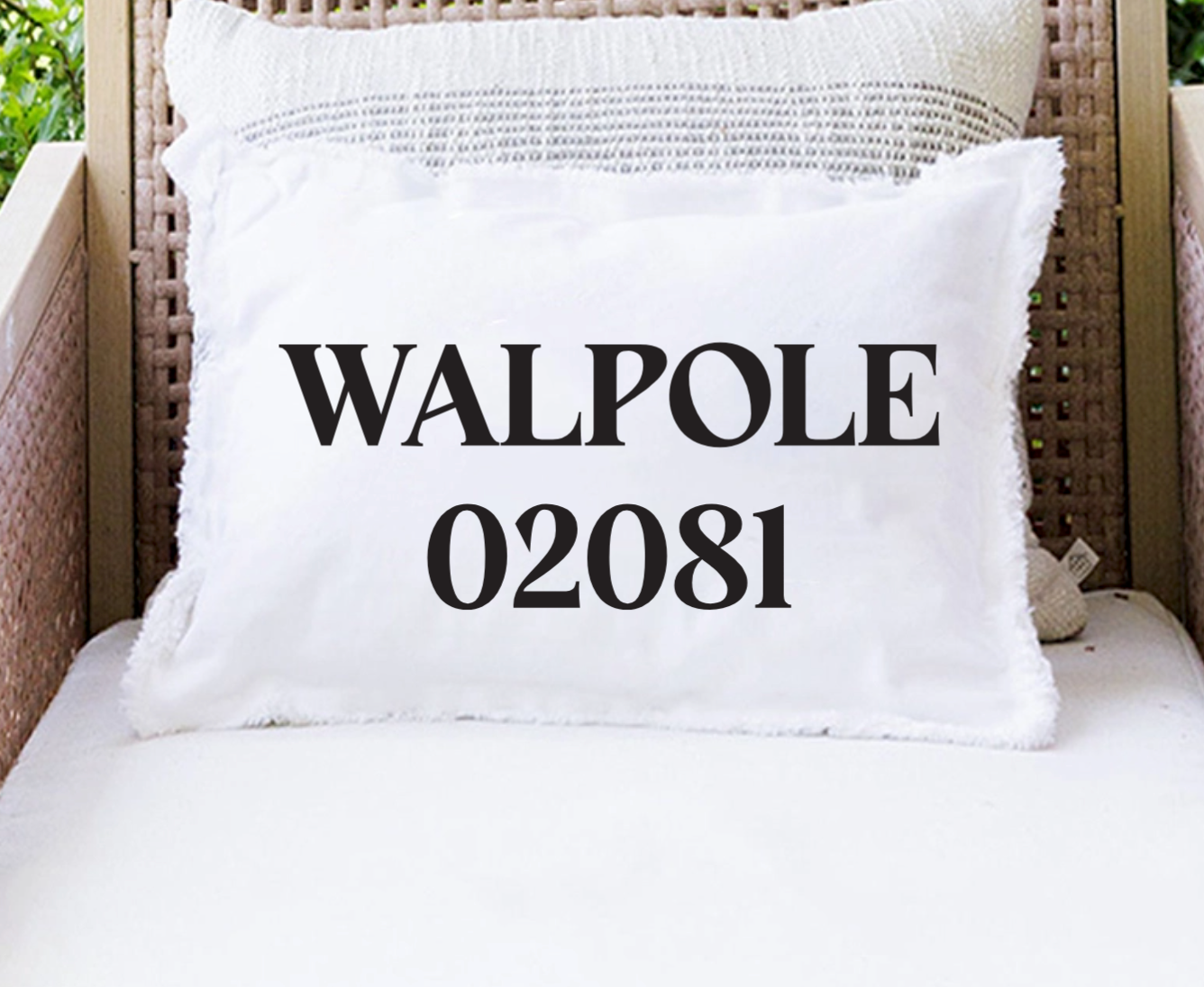 A white rectangular throw pillow with fringed edges sits on a white cushion in a wicker chair. The front of the pillow is printed with the text 'WALPOLE 02081' in black serif font. Available at Be Charmed