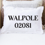 A white rectangular throw pillow with fringed edges sits on a white cushion in a wicker chair. The front of the pillow is printed with the text 'WALPOLE 02081' in black serif font. Available at Be Charmed