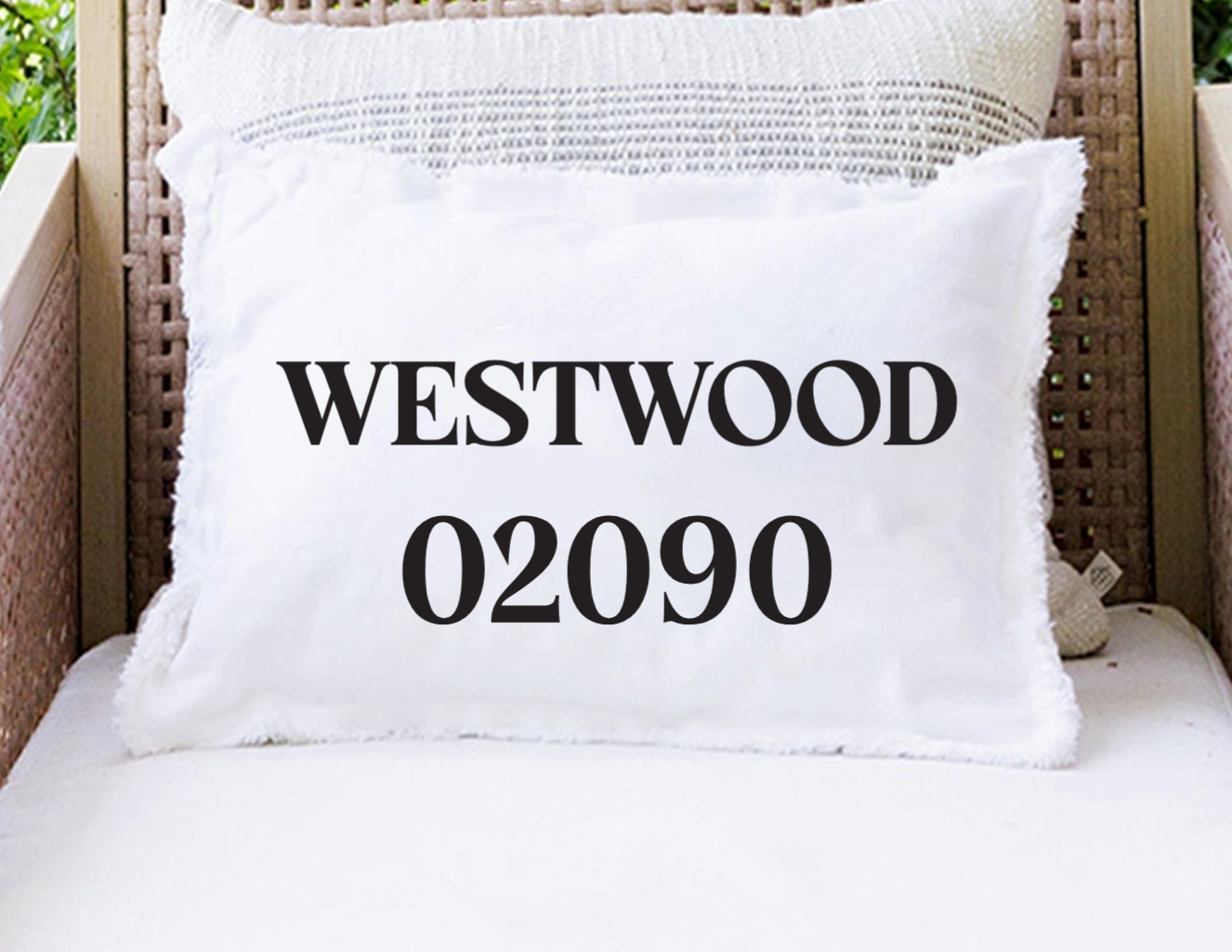 A white rectangular throw pillow with fringed edges sits on a white cushion in a wicker chair. The front of the pillow is printed with the text 'WESTWOOD 02090' in large black serif letters. Available at Be Charmed