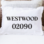 A white rectangular throw pillow with fringed edges sits on a white cushion in a wicker chair. The front of the pillow is printed with the text 'WESTWOOD 02090' in large black serif letters. Available at Be Charmed