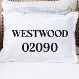 A white rectangular throw pillow with fringed edges sits on a white cushion in a wicker chair. The front of the pillow is printed with the text 'WESTWOOD 02090' in large black serif letters. Available at Be Charmed
