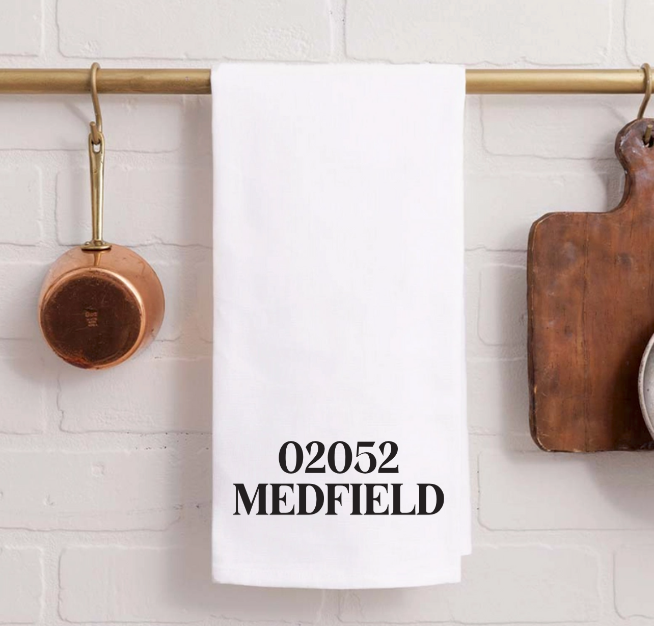 A Rustic Marlin white kitchen towel with the text '02052 MEDFIELD' printed in black, hanging from a brass rod on a white brick wall with kitchen props. Available at Be Charmed