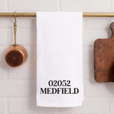 A Rustic Marlin white kitchen towel with the text '02052 MEDFIELD' printed in black, hanging from a brass rod on a white brick wall with kitchen props. Available at Be Charmed