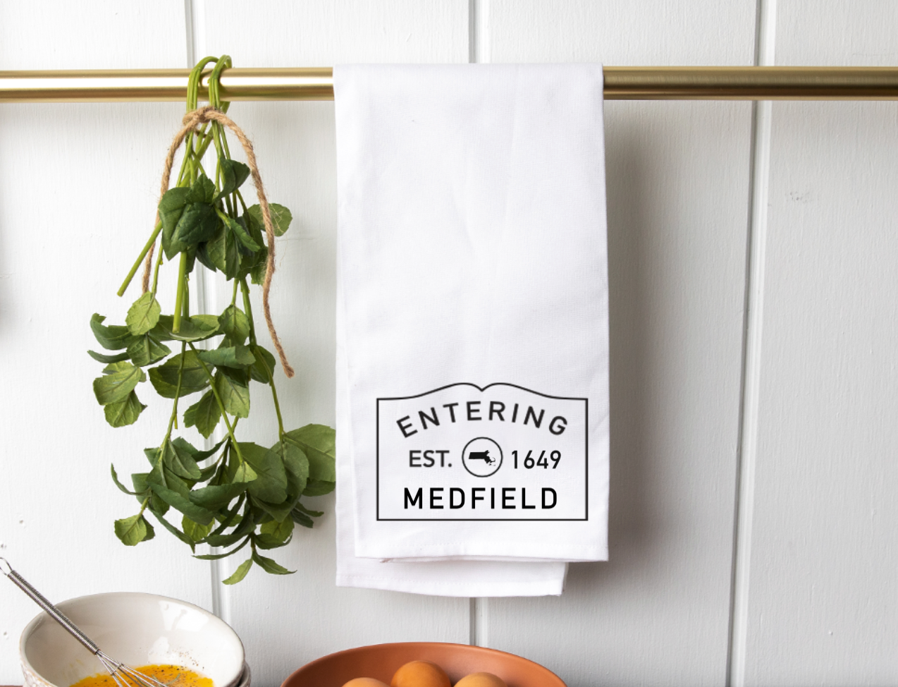 A Rustic Marlin tea towel with vintage-style 'ENTERING EST. 1649 MEDFIELD' stamp graphic hangs from a brass rod next to a bunch of fresh green herbs. Available at Be Charmed