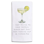 A Rustic Marlin white kitchen towel featuring a printed illustration of a margarita with a lime slice and the humorous text, 'I KEEP TRYING TO MAKE PROTEIN SHAKES BUT THEY KEEP TURNING OUT AS MARGARITAS'. Available at Be Charmed