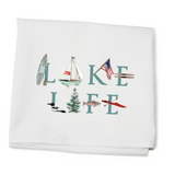 A white cotton kitchen towel with 'Lake Life' text and an illustration that includes a sailboat, a fish, a pine tree, and a red bird. Available at Be Charmed