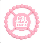 A pink silicone baby teether with a circular beaded outer ring and a central round piece that reads 'let's take a selfie' in white text, with 'BELLA TUNNO' printed below. Available at Be Charmed