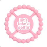 A pink silicone baby teether with a circular beaded outer ring and a central round piece that reads 'let's take a selfie' in white text, with 'BELLA TUNNO' printed below. Available at Be Charmed