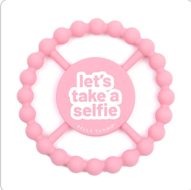 A pink silicone baby teether with a circular beaded outer ring and a central round piece that reads 'let's take a selfie' in white text, with 'BELLA TUNNO' printed below. Available at Be Charmed