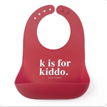 A red Bella Tunno silicone baby bib with a front pocket and the printed text 'k is for kiddo.' in white, on a plain white background. Available at Be Charmed