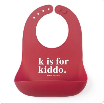 A red Bella Tunno silicone baby bib with a front pocket and the printed text 'k is for kiddo.' in white, on a plain white background. Available at Be Charmed