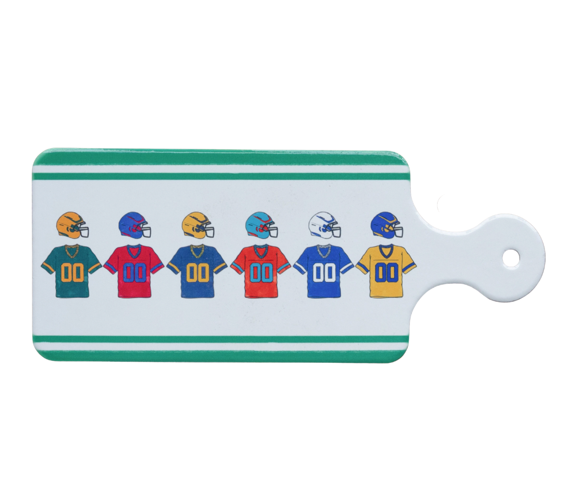 A rectangular Boston International white cutting board or decorative board with a handle, featuring a row of colorful football helmets and jerseys printed on the front. The board has green stripes at the top and bottom and a white handle with a hole. Available at Be Charmed
