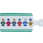 A rectangular Boston International white cutting board or decorative board with a handle, featuring a row of colorful football helmets and jerseys printed on the front. The board has green stripes at the top and bottom and a white handle with a hole. Available at Be Charmed